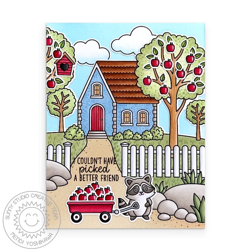 Raccoon pulling a wagon with apple trees in front of a blue house with a white picket fence, handmade fall scene card.