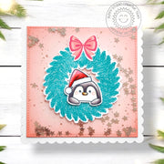 Handmade Christmas confetti shaker card featuring a penguin in a teal holiday wreath, with a pink bow on a peach background.