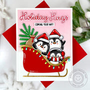 Chubby penguins, tree, and gifts piled in a red sleigh decorated with colorful lights, "Holiday Hugs" Christmas card.